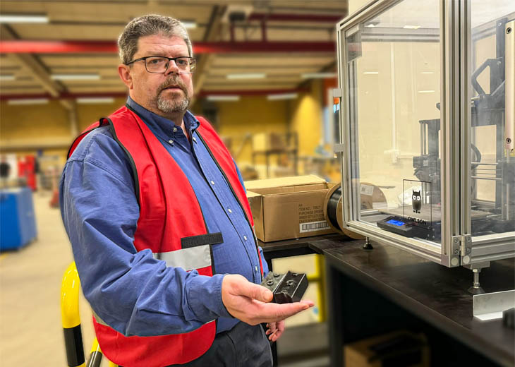 Peter stands beside a line of state-of-the-art 3D printers. In his hand, he proudly holds a small, yet crucial, 3D printed safety component, showcasing the company's commitment to precision and safety in every detail.