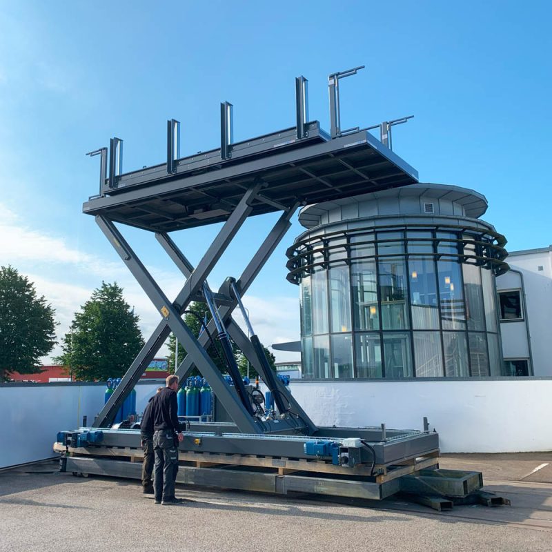 Two technicians examine a tall railbound lift platform in front of SIGI Europe’s curved glass office building.