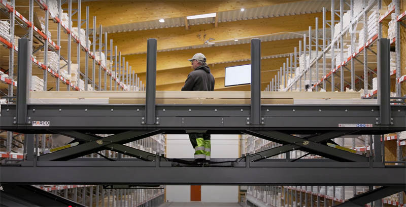 Man standing on an elevated platform lift between high warehouse racks filled with wood profiles.